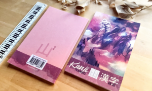 Deux cahiers d'entraînement aux kanji sont posés sur une table en bois, à côté d'une règle et de feuilles séchées. L'un des cahiers a une couverture rose avec un texte en japonais ; l'autre représente un paysage de montagne et porte la mention « Kanji Training » sur la couverture.