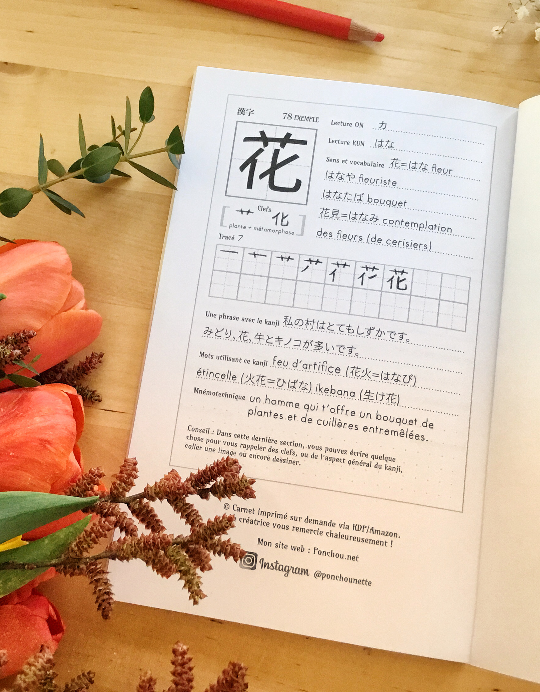 Un cahier d'exercices de japonais ouvert présente le kanji signifiant « fleur » (花), accompagné d'explications en français, de l'ordre des traits et d'un espace pour s'entraîner. Des tulipes orange et des feuilles vertes sont posées sur une table en bois clair, à côté du livre.
