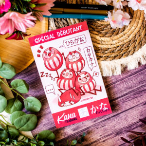 Un livret « Kana Training かな - Cahier d’écriture japonaise - Spécial Débutant » aux couleurs vives, mettant en scène des personnages de daruma de bande dessinée, accompagnés de fleurs, de feuilles vertes, d’un stylo en bois et d’un tapis posé sur une surface en bois. On y distingue à la fois l’écriture japonaise et le texte français « Spécial Débutant ».