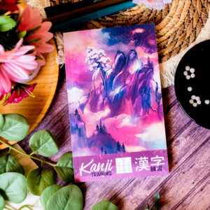 Le livre « Kanji Training 漢字 Grue 鶴 : Cahier d’entraînement à l’écriture japonaise », dont la couverture colorée représente un paysage de montagne, repose sur une table en bois au milieu de fleurs, de feuilles vertes, de stylos, d’un plat noir contenant des ardoises blanches et d’un set de table tissé.
