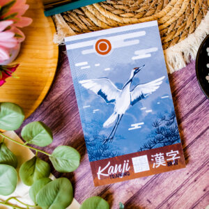L’ouvrage « Kanji Training 漢字 Grue 鶴 : Cahier d’entraînement à l’écriture japonaise », dont la couverture est ornée d’une illustration représentant une grue en plein vol, est présenté sur un support en bois, entouré de feuilles, d’une fleur et d’une natte tressée.