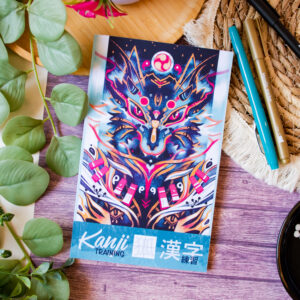 Le livre « Kanji Training 漢字 Grue 鶴 : Cahier d’entraînement à l’écriture japonaise », dont la couverture est ornée d’un motif abstrait et coloré représentant un loup, repose sur une table en bois au milieu de feuilles vertes, de stylos, d’une natte tressée et d’un petit bol rempli de pierres noires et blanches.