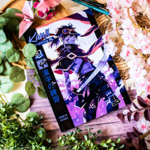 Le « Kanji Training 漢字 Grue 鶴 : Cahier d’entraînement à l’écriture japonaise » repose sur une surface en bois ; sa couverture illustrée représentant un ninja est entourée de fleurs roses et blanches, de feuilles vertes et de stylos bleus.