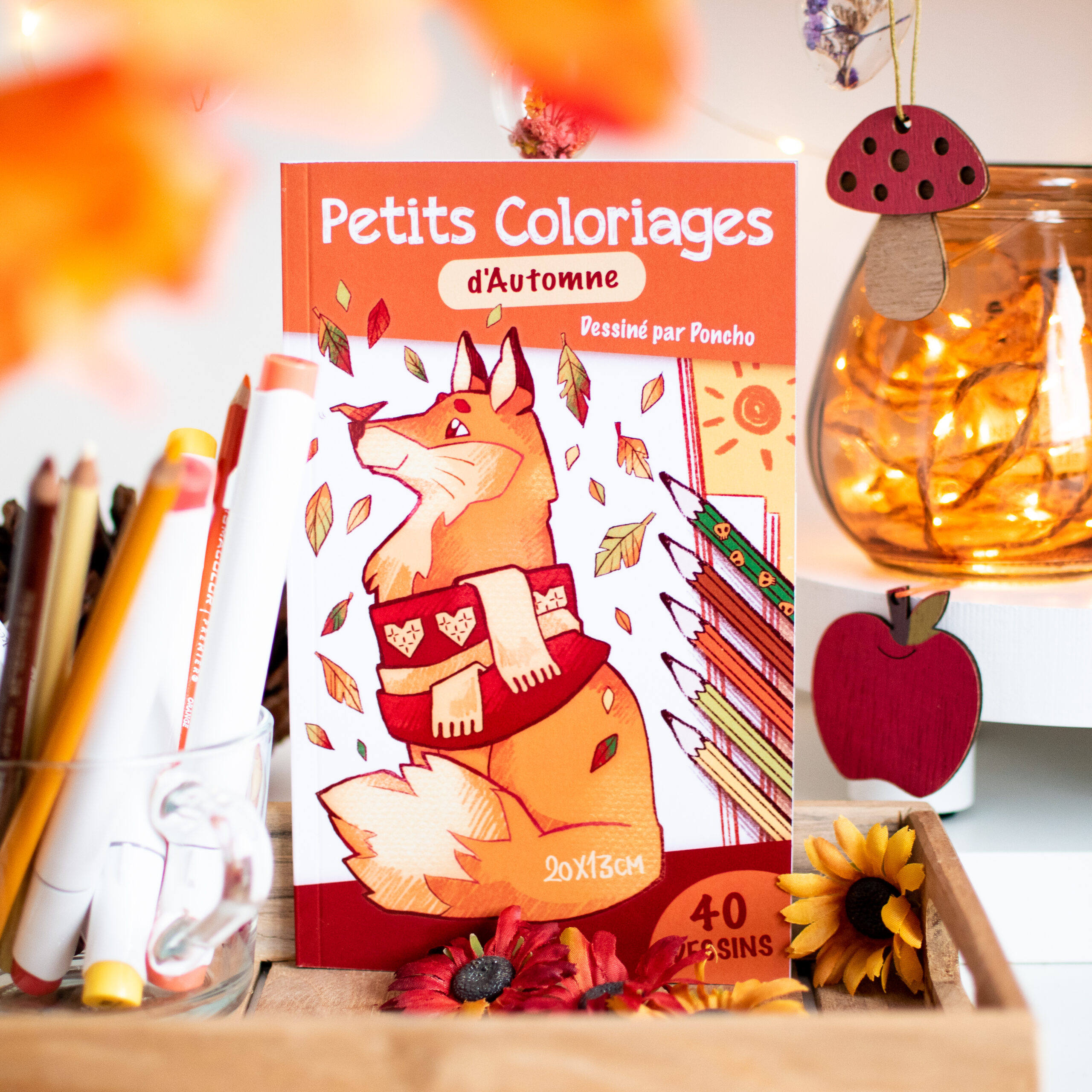 Le livre « Petits Coloriages d'Automne », dont la couverture représente un renard portant une écharpe, est exposé au milieu d'un décor sur le thème de l'automne, de feutres de couleur et d'un bocal rempli de guirlandes lumineuses orange.