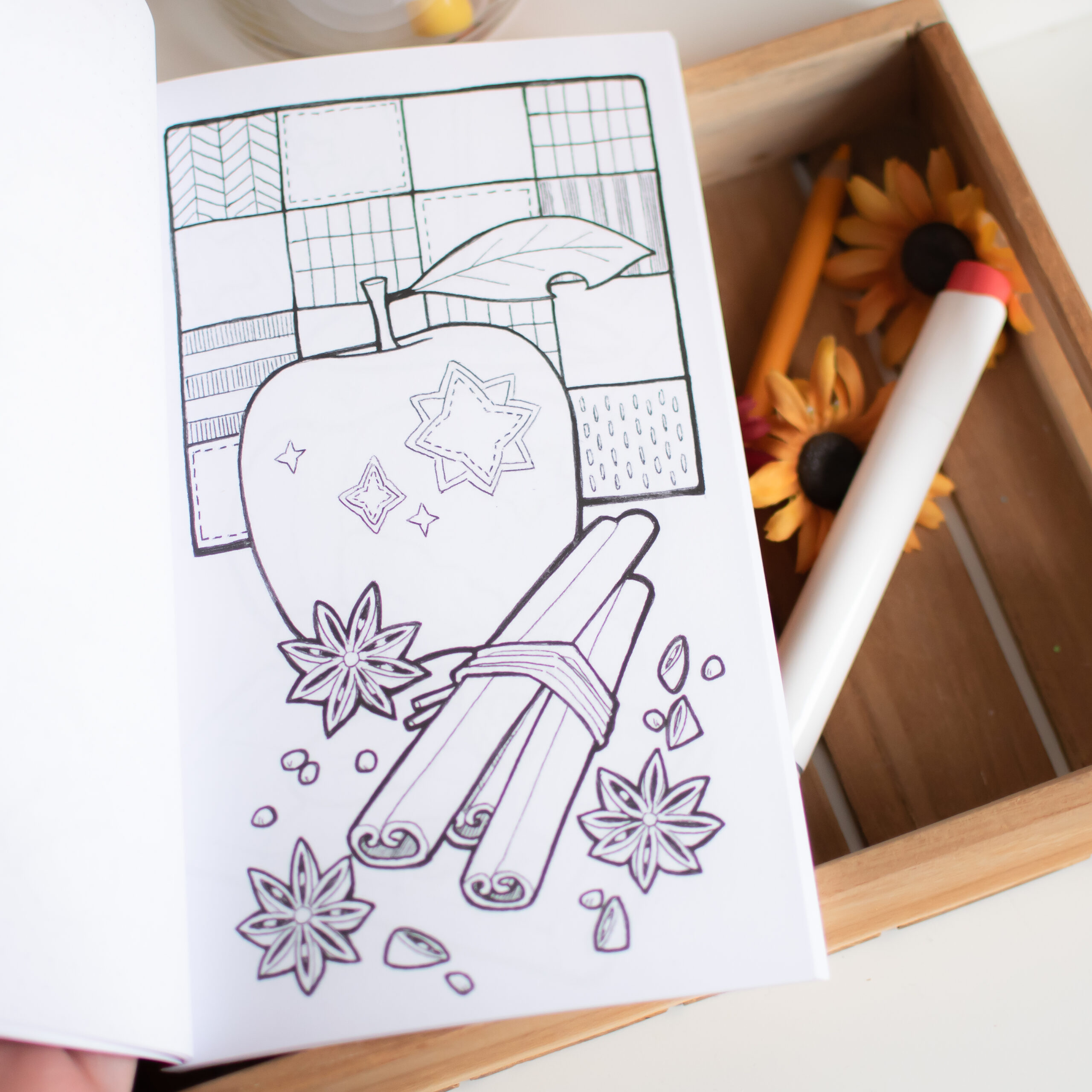 Petits Coloriages d'Automne : une page de coloriage représentant une pomme, des bâtons de cannelle, de l'anis étoilé et des motifs géométriques. À côté se trouve une boîte en bois contenant des crayons, un feutre blanc et des tournesols artificiels.