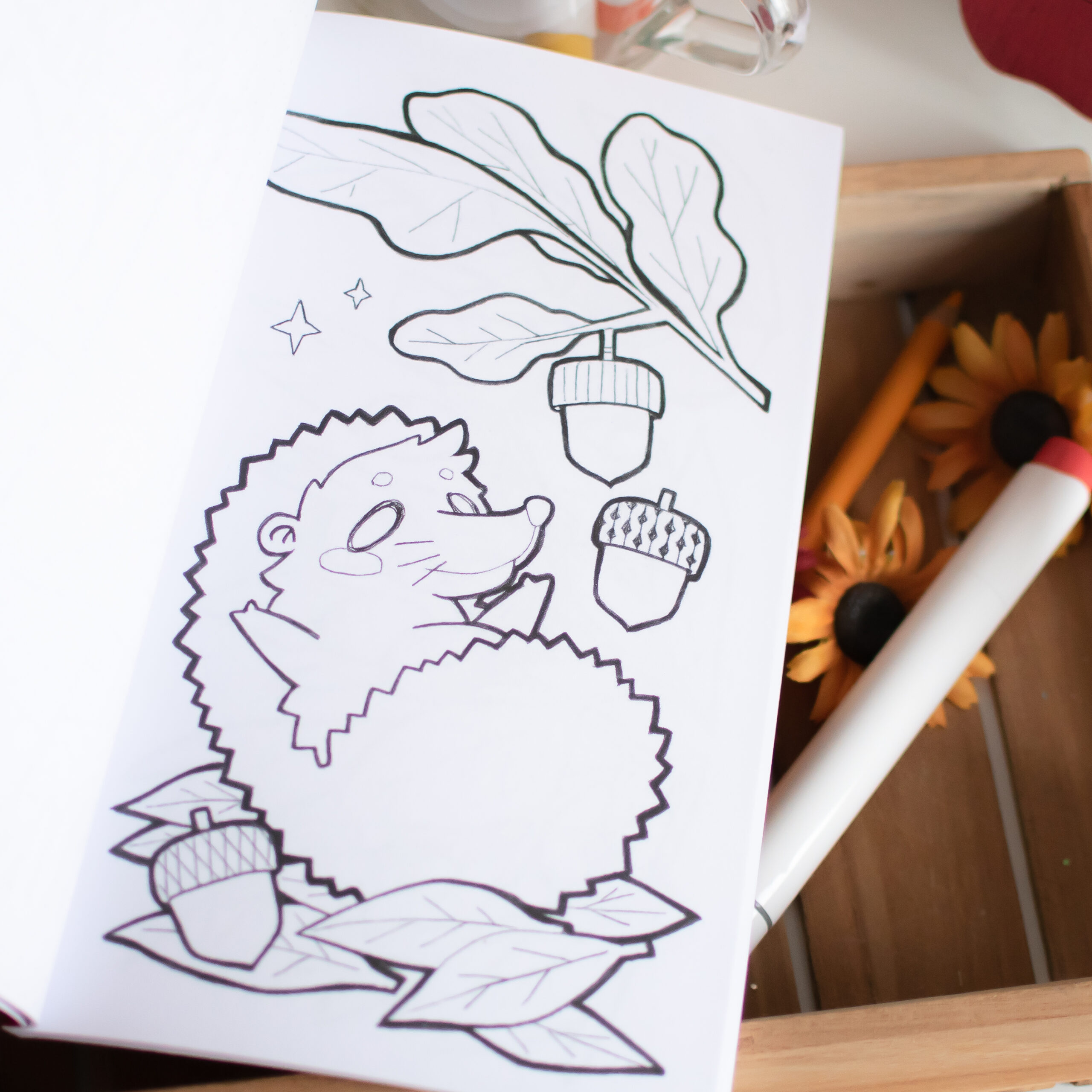 « Petits Coloriages d'Automne » est une page de coloriage en noir et blanc représentant un hérisson, des feuilles, des glands et des étoiles, disposés sur une boîte en bois à côté de tournesols et d'outils de coloriage tels qu'un crayon orange et un feutre rouge et noir.