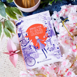 Le livre « Rendez-vous au Soleil couchant » est présenté sur un fond décoratif orné de fleurs roses, de feuilles vertes, d'une grue en papier rose et d'un bol rempli de bâtonnets en bois. Sa couverture est agrémentée d'illustrations fantaisistes dans les tons violets et orange.