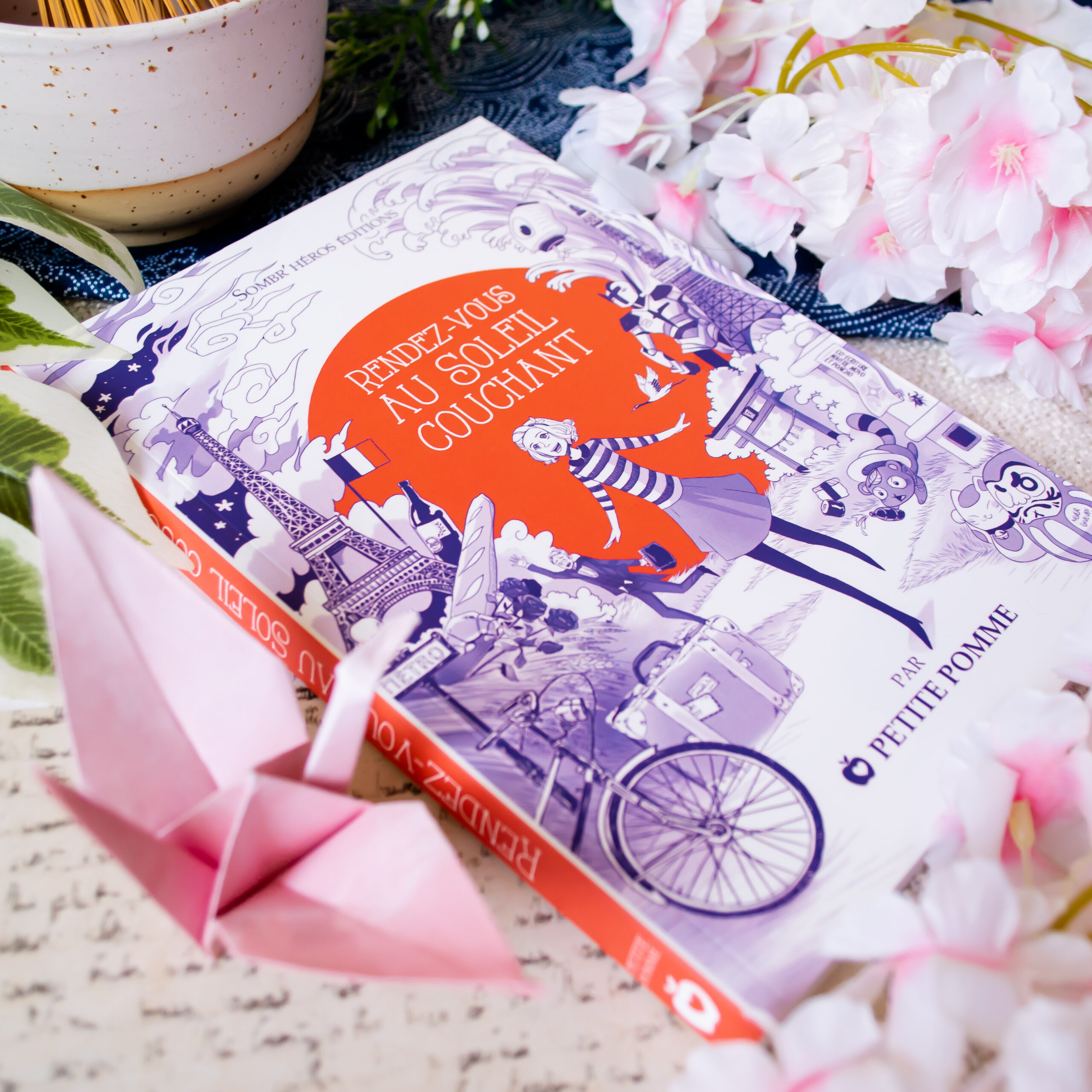Le livre français *Rendez-vous au Soleil couchant* repose sur une table, entouré de fleurs roses, d'une grue en origami rose et de verdure. Sa couverture est ornée d'illustrations fantaisistes dans les tons violets et orange.