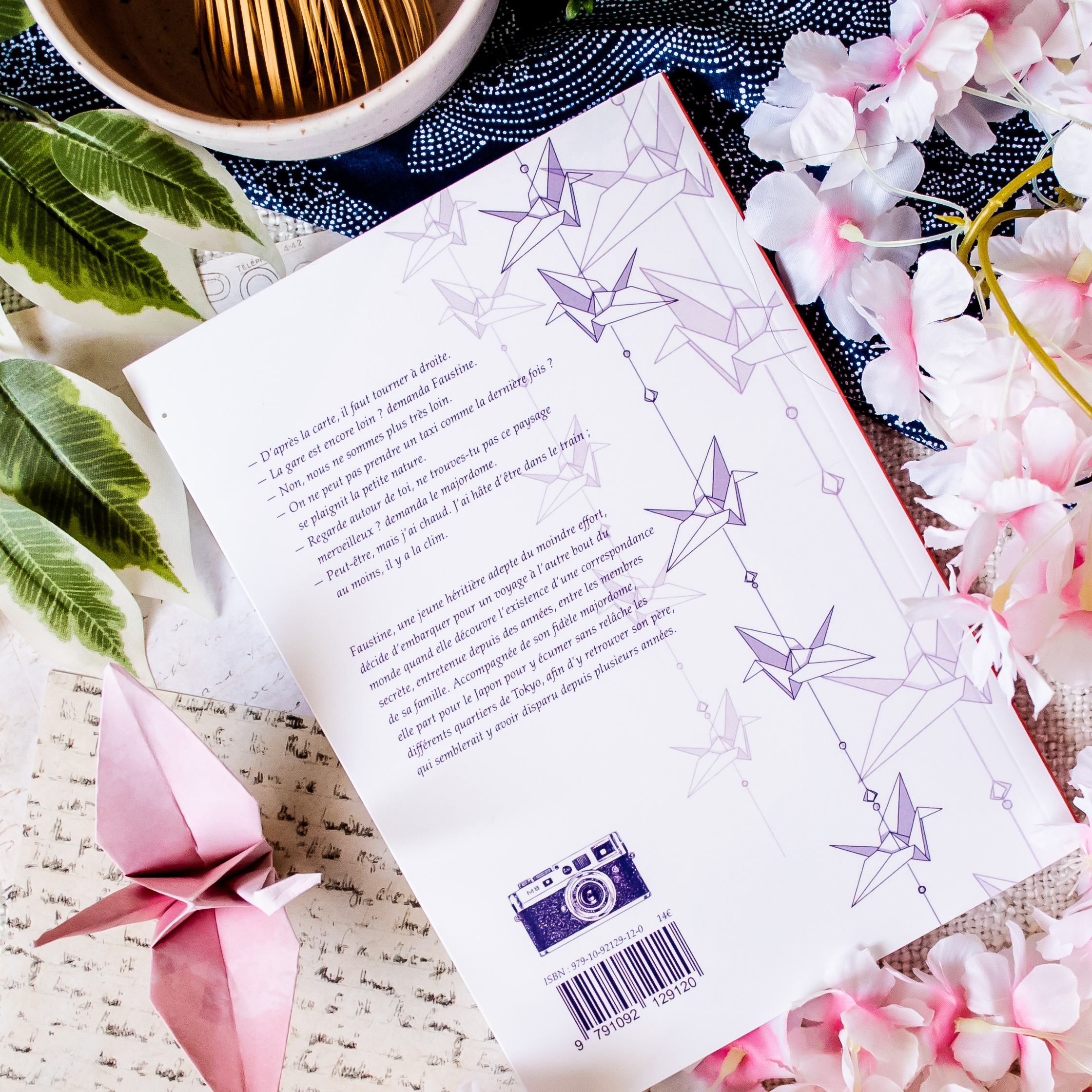 Le livre « Rendez-vous au Soleil couchant » repose sur une table ornée de fleurs roses, de feuilles vertes, d'une lettre manuscrite, d'une grue en origami rose et d'un bol rempli de bâtonnets en bois. Des grues en origami sont pliées sur sa quatrième de couverture.