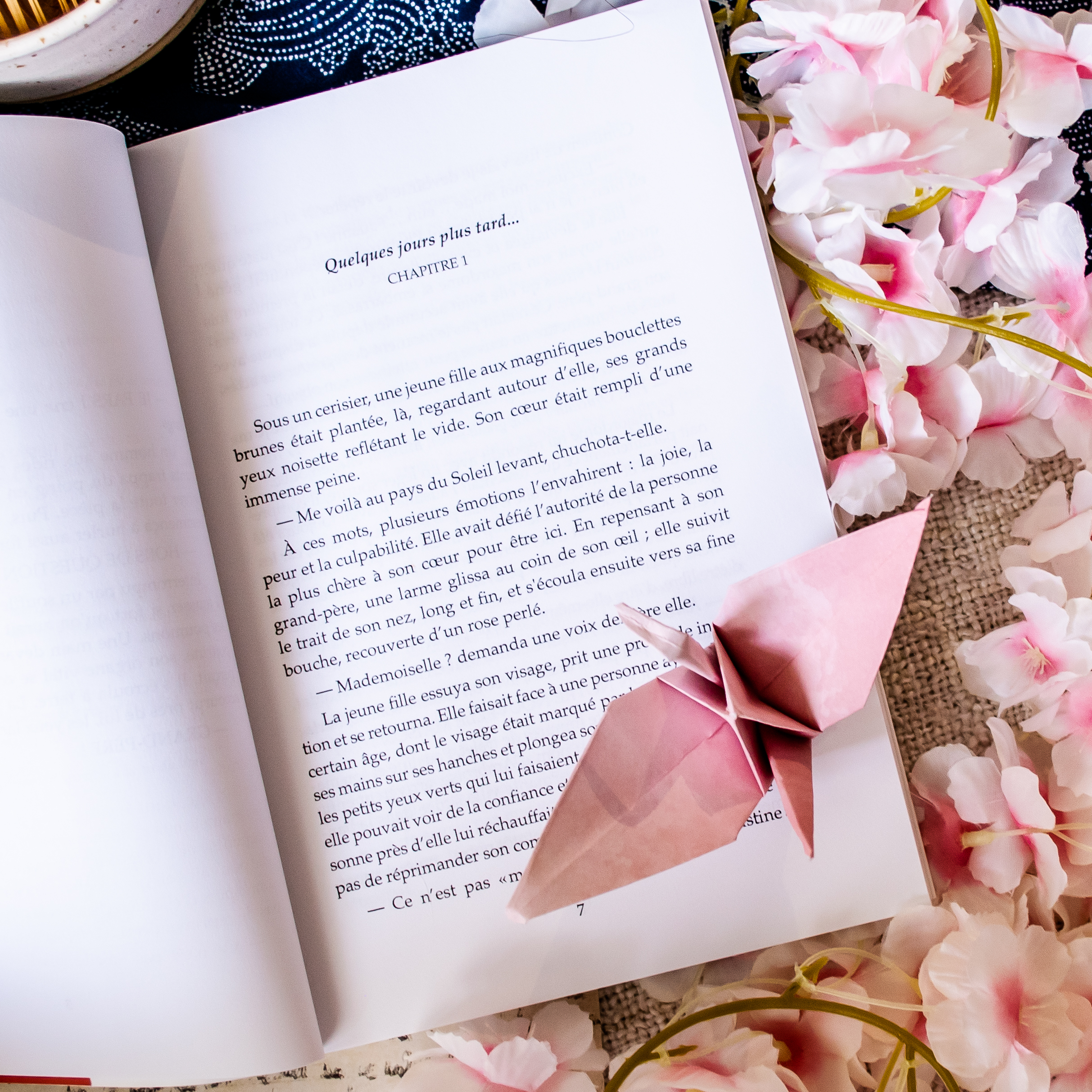 Un exemplaire de *Rendez-vous au Soleil couchant* est ouvert sur une surface, entouré de fleurs de cerisier roses, avec une grue en origami rose posée sur ses pages.