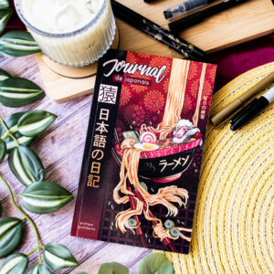 Le « Journal de japonais » 日記 Carnet de bord 猿 Saru, dont la couverture est ornée d'une illustration représentant des ramen, repose sur une table entourée de feuilles vertes, d'une bougie, de stylos et d'un chapeau de paille.