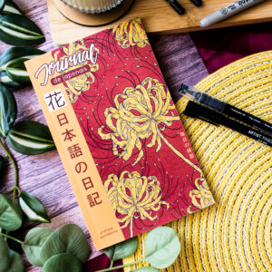 Le « Journal de japonais » 日記 Carnet de bord 猿 Saru, dont la couverture rouge est ornée de motifs floraux jaunes, repose sur une natte tressée, au milieu de feuilles vertes, de stylos, d'un plateau en bambou et de divers autres articles de papeterie.