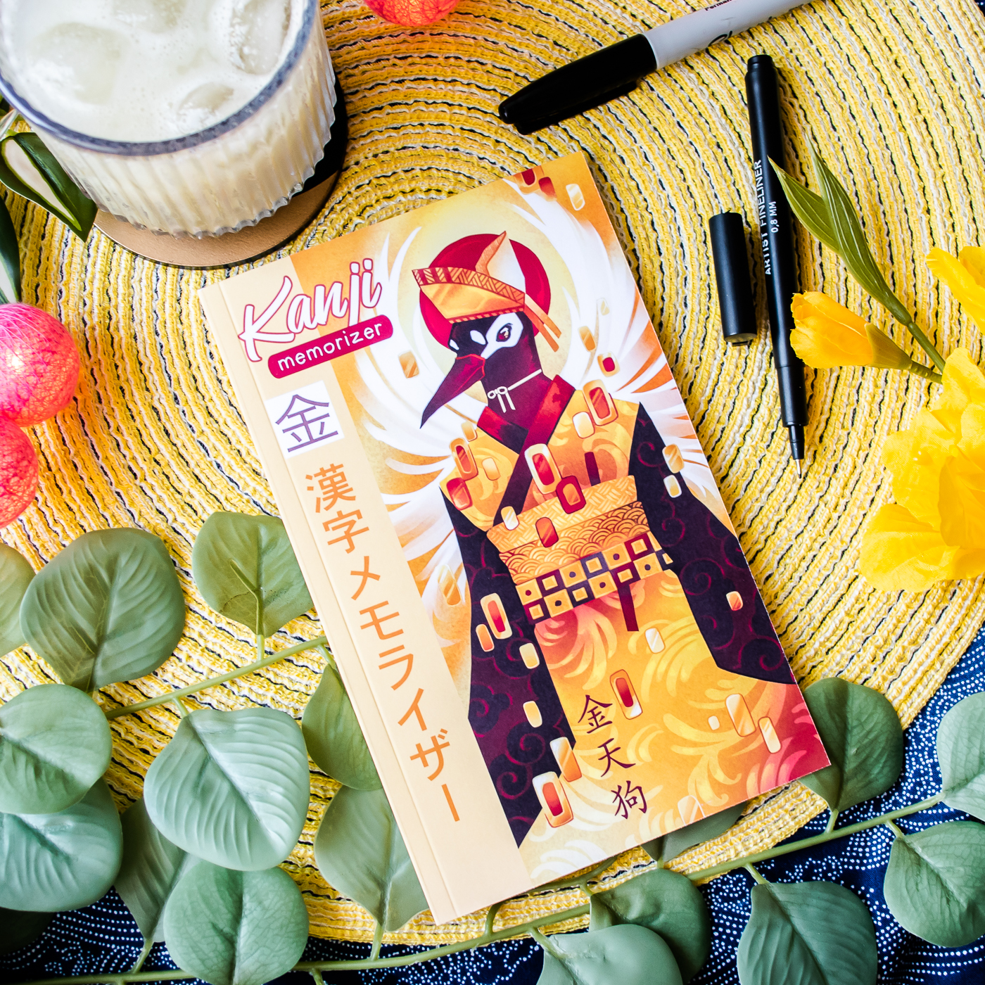 Un exemplaire de « Kanji Memorizer 学生 Étudiant : Répertoire mnémotechnique kanji à remplir », dont la couverture aux couleurs vives est ornée d’une grue stylisée, repose sur un tapis tissé jaune, au milieu de feuilles vertes, d’un assortiment de stylos, d’une boisson et d’éléments décoratifs orange et jaunes.