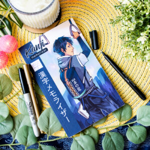 Un livre de style manga intitulé « Kanji Memorizer 学生 Étudiant : Répertoire mnémotechnique kanji à remplir », contenant du texte en japonais, repose sur des feuilles et est entouré de stylos, d’un cahier et d’une boisson. La couverture représente un garçon en train de dessiner des caractères kanji.