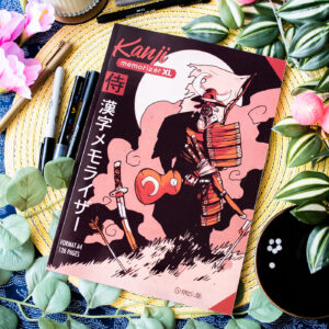 Le cahier d'exercices « Kanji Memorizer 学生 Étudiant : Répertoire mnémotechnique kanji à remplir », dont la couverture est ornée d'une illustration représentant un samouraï et d'un texte en japonais, repose sur une natte tissée, au milieu de stylos, de feuilles et de fleurs.