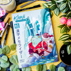 Un cahier d’exercices aux couleurs vives intitulé « Kanji Memorizer 学生 Étudiant : Répertoire mnémotechnique kanji à remplir », dont la couverture représente une grenouille en tenue de samouraï, repose parmi des feuilles, des stylos, des fruits et une bougie. Ce dessin coloré est entouré de matériel artistique et d’objets décoratifs.