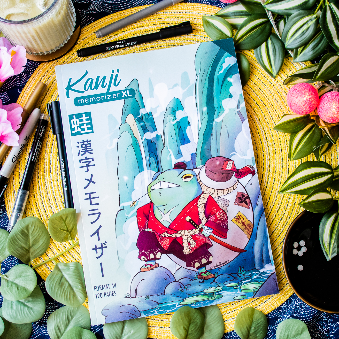 Un cahier d’exercices aux couleurs vives intitulé « Kanji Memorizer 学生 Étudiant : Répertoire mnémotechnique kanji à remplir », dont la couverture représente une grenouille en tenue de samouraï, repose parmi des feuilles, des stylos, des fruits et une bougie. Ce dessin coloré est entouré de matériel artistique et d’objets décoratifs.
