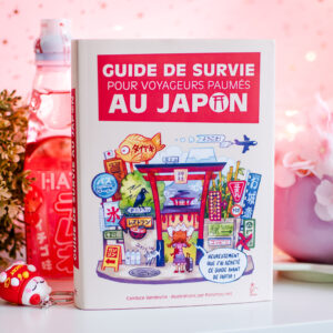 Le « Guide de survie pour voyageurs perdus au Japon » est posé sur une table rose, entouré de snacks japonais, avec en arrière-plan des décorations représentant des fleurs de cerisier.