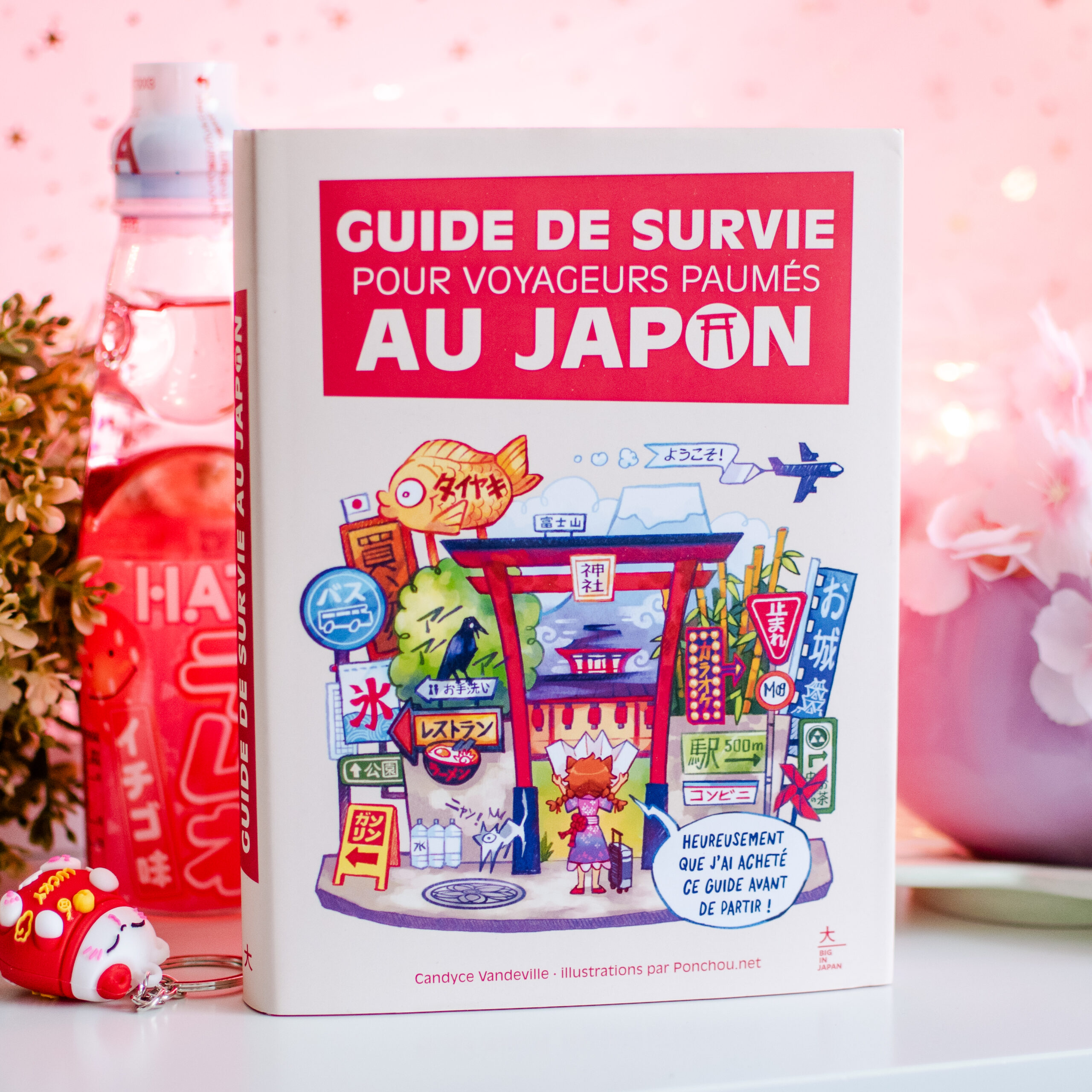 Le « Guide de survie pour voyageurs perdus au Japon » est posé sur une table rose, entouré de snacks japonais, avec en arrière-plan des décorations représentant des fleurs de cerisier.