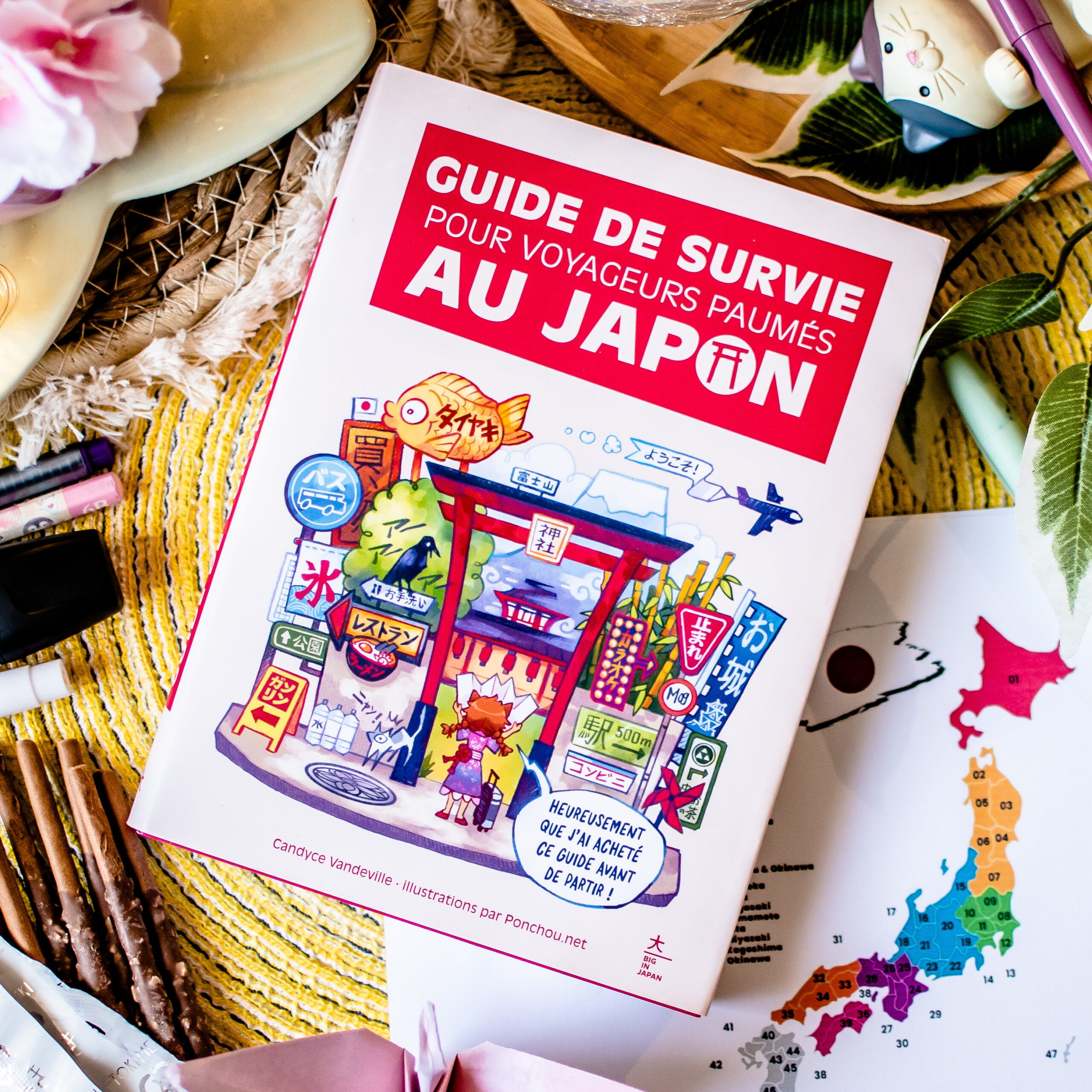 Un exemplaire du « Guide de survie pour voyageurs paumés au Japon » est posé sur une table, entouré d’objets colorés, notamment des stylos, une plante et une carte partiellement visible sur laquelle figurent des régions numérotées. La couverture du livre présente des panneaux japonais illustrés et une porte de sanctuaire rouge.