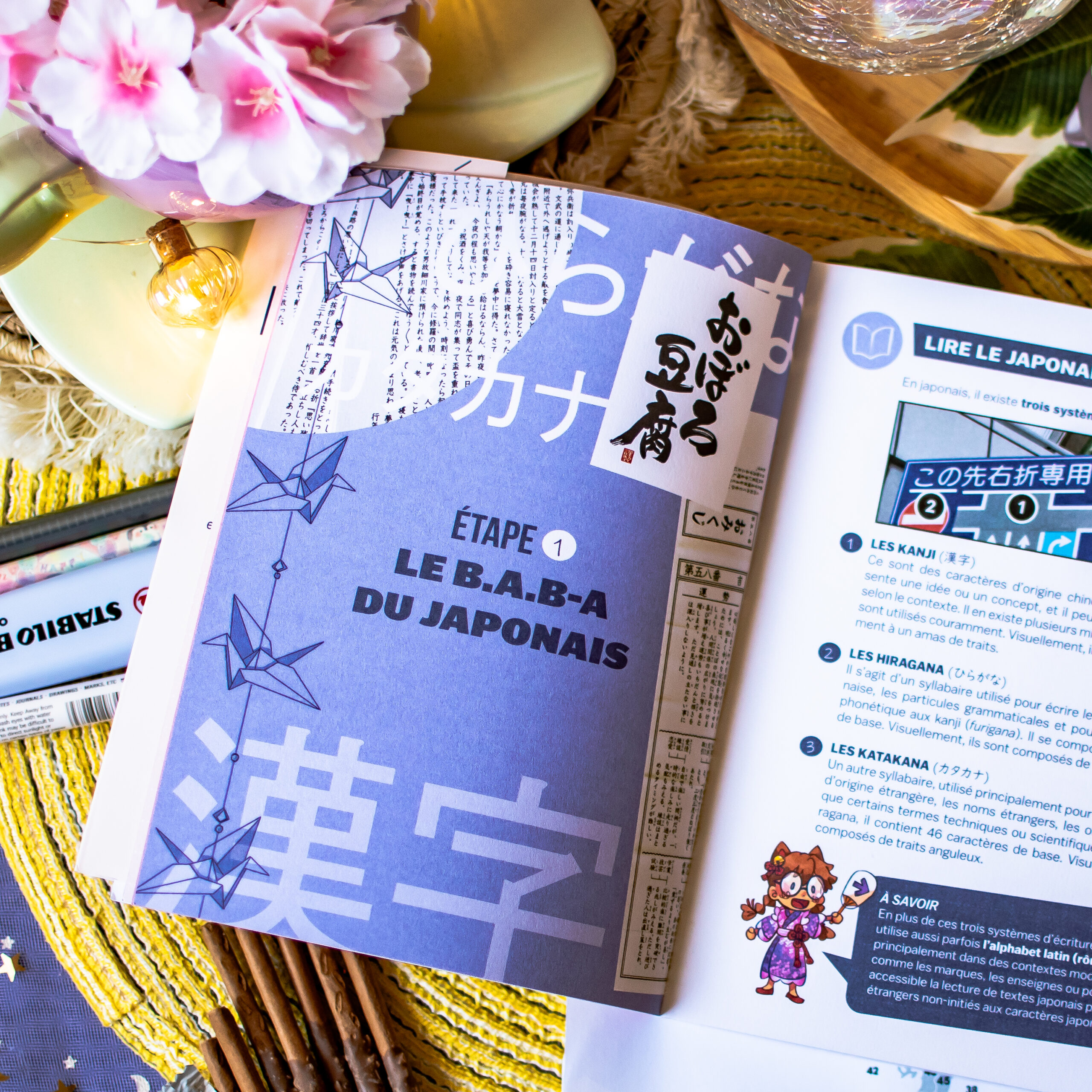 Un exemplaire ouvert du « Guide de survie pour voyageurs paumés au Japon » repose sur une table, à côté d’objets décoratifs, d’un stylo et d’une fleur. La page visible comporte du texte en français et en japonais, des grues en origami et des illustrations décoratives.