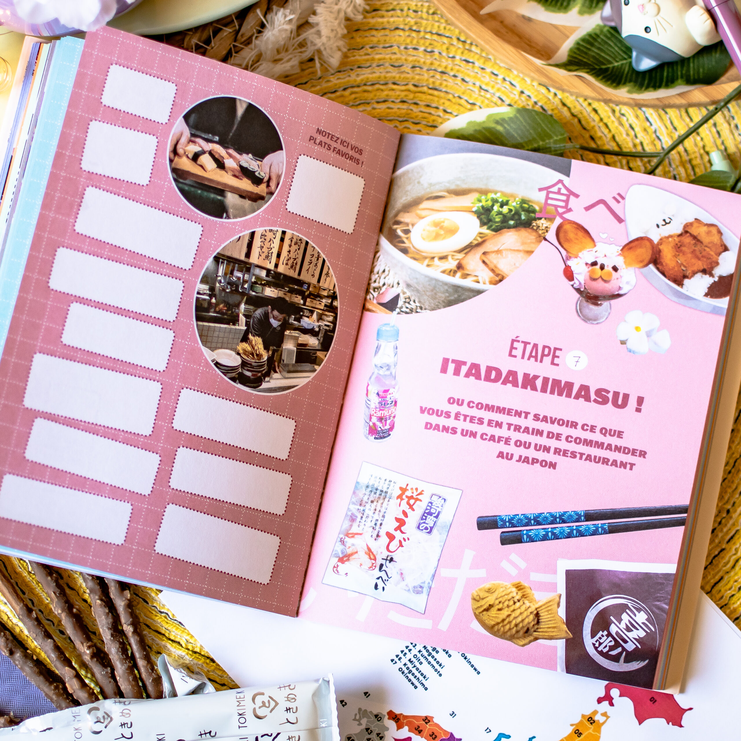 Un exemplaire ouvert du « Guide de survie pour voyageurs paumés au Japon » présente une page consacrée à la gastronomie, avec des photos de ramen, de boissons et de snacks, un texte en français expliquant comment commander au Japon, ainsi qu’un calendrier vierge. Divers snacks et articles de papeterie sont disposés sur une natte tressée autour du guide.