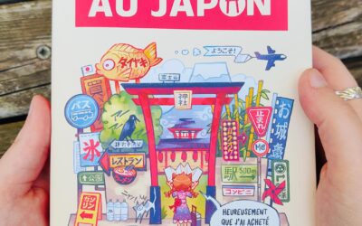 Sur une surface en bois, on voit quelqu'un tenant le livre en français « Guide de survie pour voyageur paumé au Japon », dont la couverture est ornée d'illustrations colorées représentant des plats japonais, des panneaux et des sites touristiques.