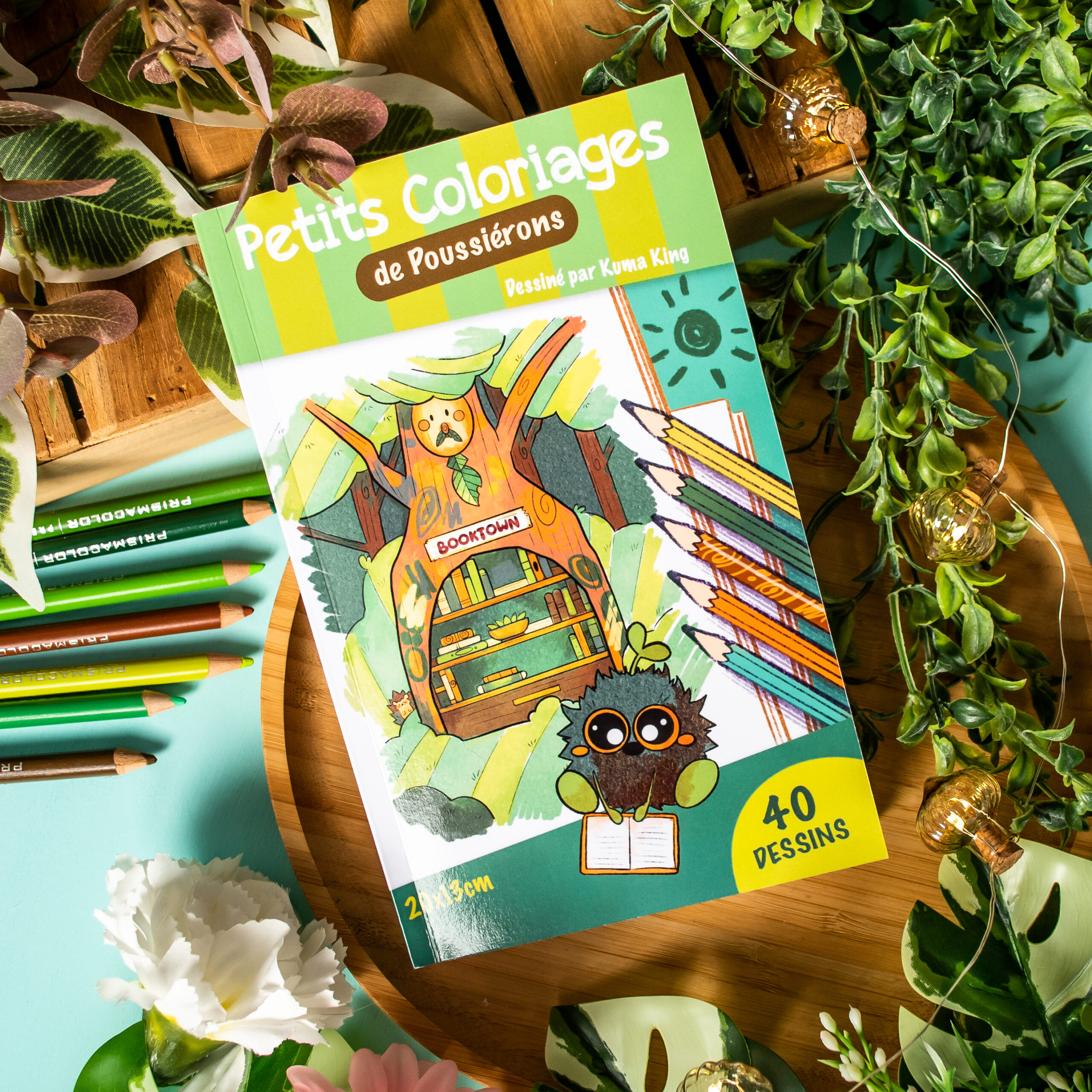Le livre de coloriage français « Coloriages de Poussiérons » repose sur un plateau en bois, entouré de plantes vertes, de fleurs et de crayons de couleur. Sa couverture représente de charmants personnages illustrés et des crayons colorés.