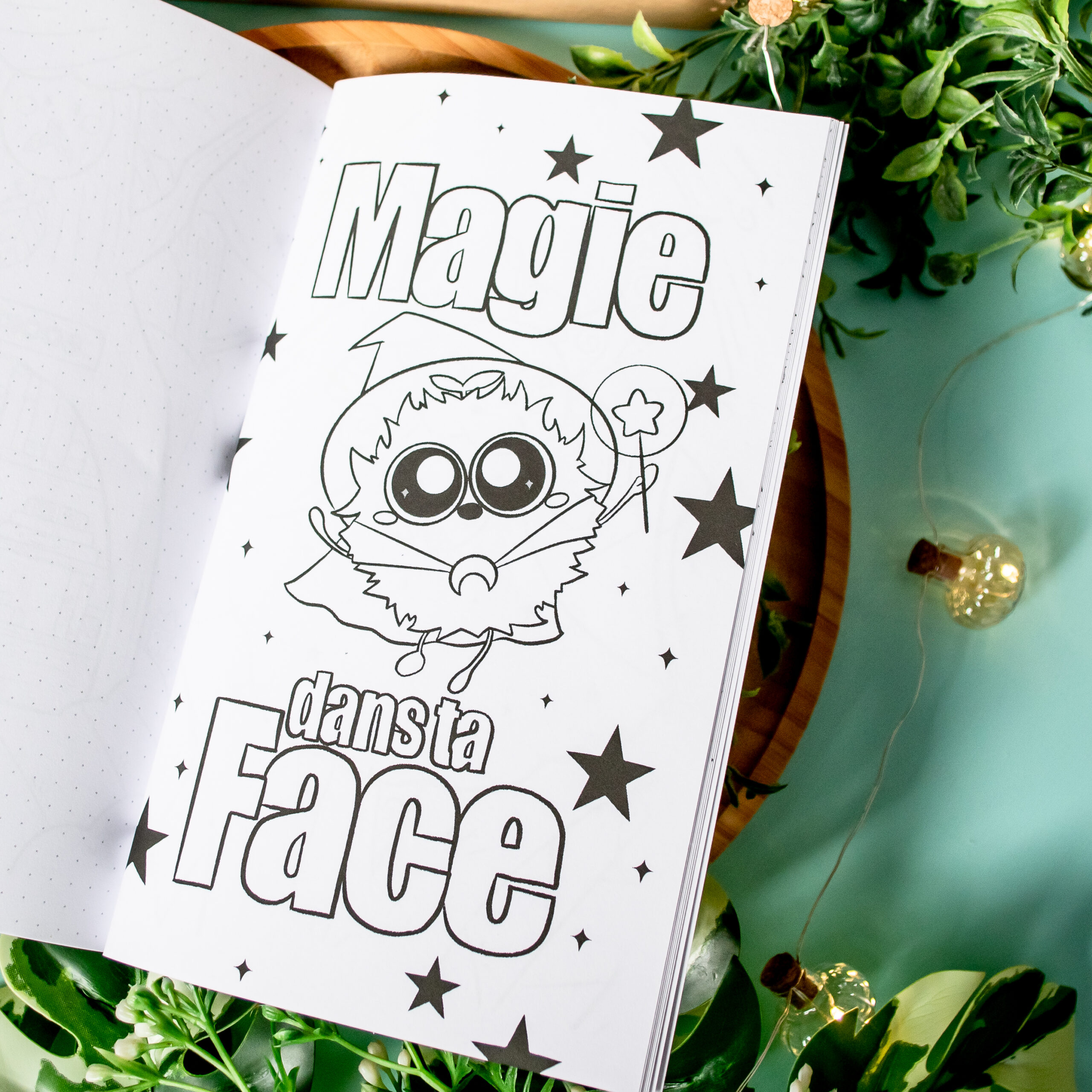 Une page du livre de coloriage « Coloriages de Poussiérons » présente le titre « Magie dans ta Face » en français ainsi qu'un sorcier de bande dessinée muni d'une baguette magique, entouré d'étoiles. Le livre ouvert repose sur une table, à côté de laquelle se trouvent des plantes et des guirlandes lumineuses.