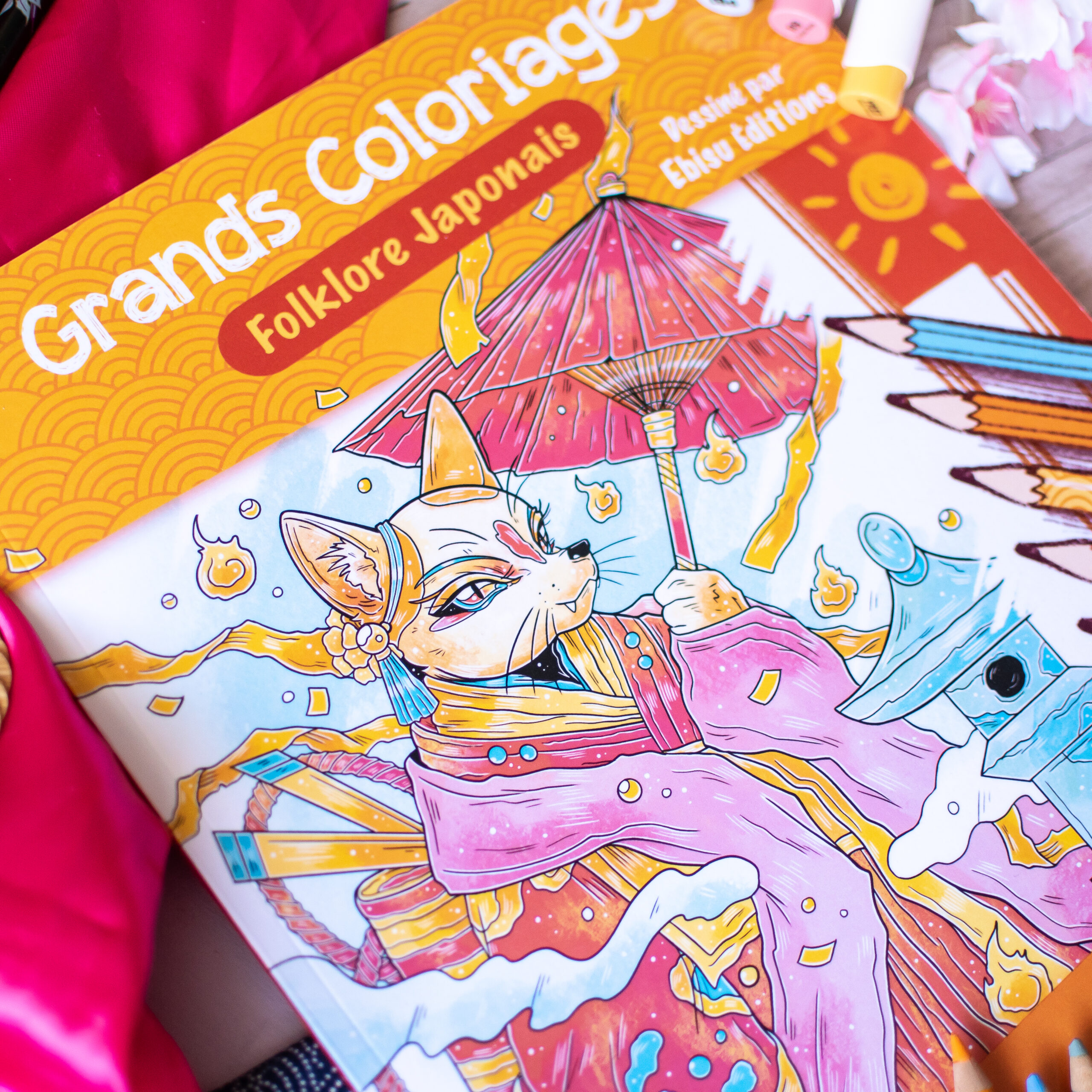 Le livre de coloriage « Coloriages Folklore Japonais » est ouvert, dévoilant une illustration détaillée représentant un renard anthropomorphe en costume traditionnel, tenant un parapluie, entouré de flammes flottantes et de motifs aux couleurs vives.