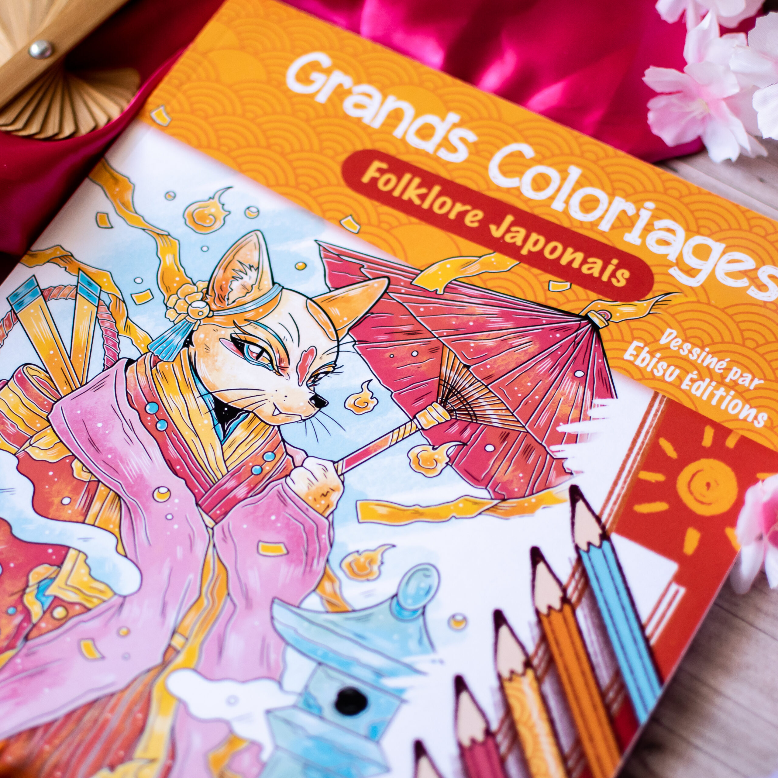 Le livre de coloriage français « Coloriages Folklore Japonais » repose sur de la soie rose ; sa couverture représente un renard en costume traditionnel japonais tenant un parapluie rouge.