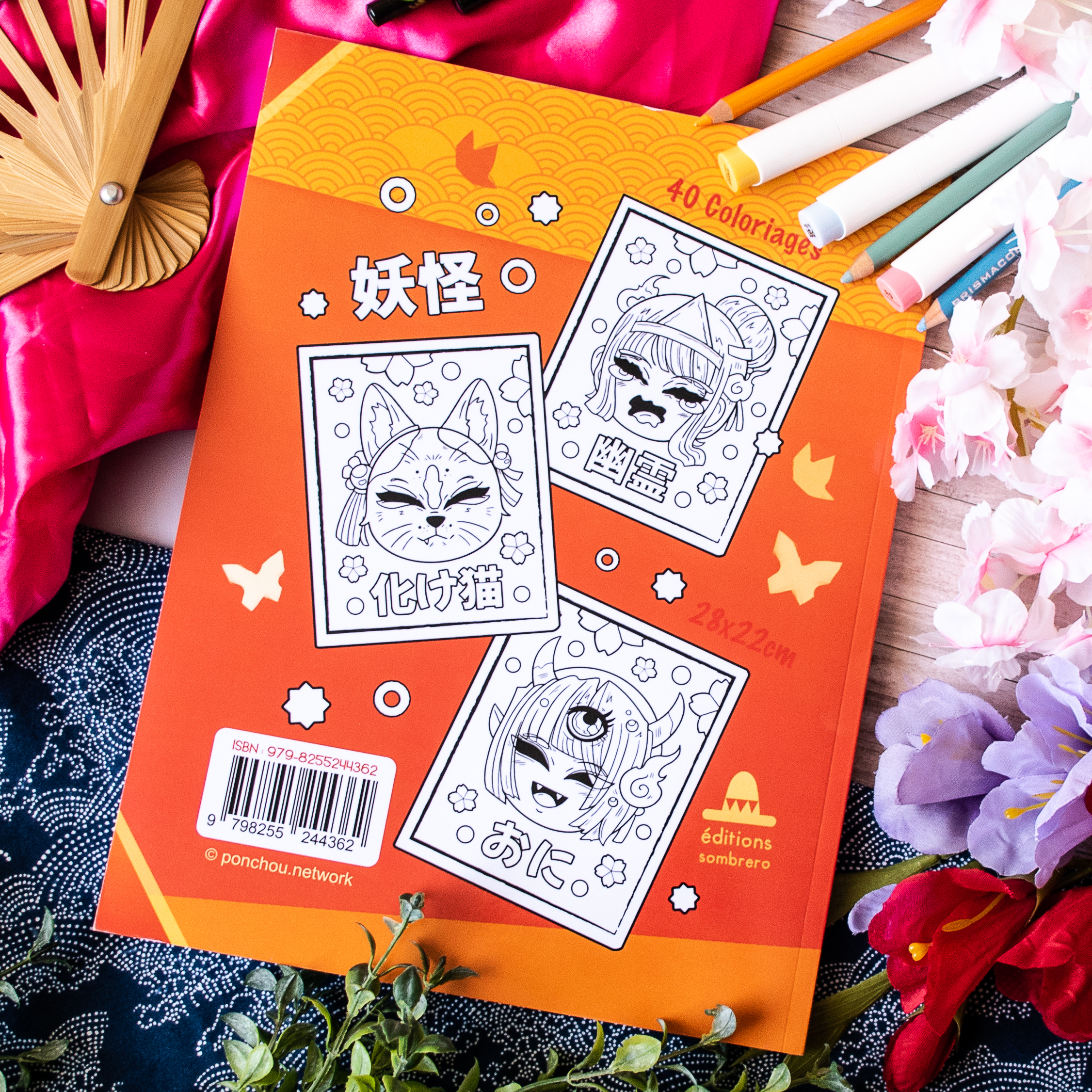 « Coloriages Folklore Japonais », un livre de coloriage orange contenant trois illustrations en noir et blanc de yokai, est présenté entouré de crayons de couleur, d'un tissu rose, d'un éventail et de fleurs roses.