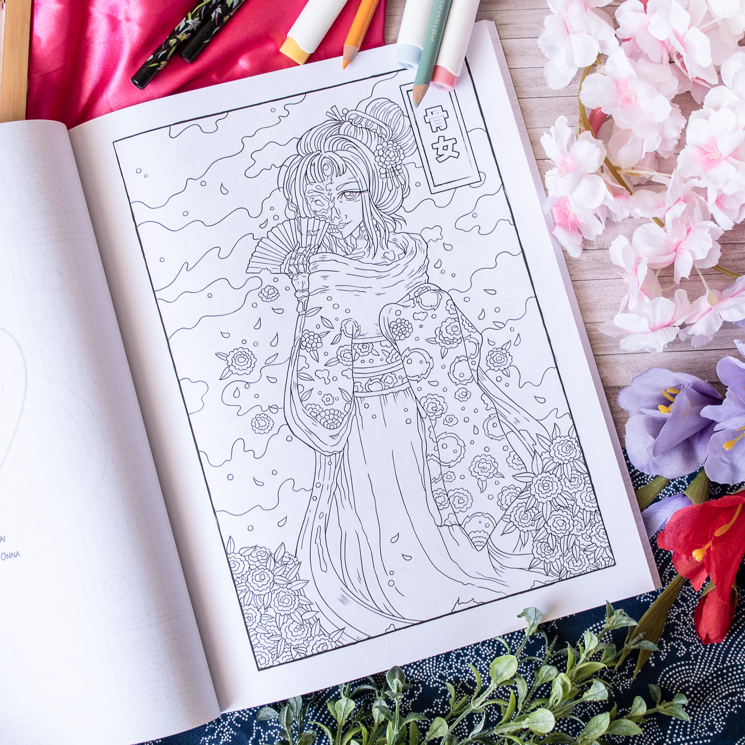 Le livre de coloriage « Coloriages Folklore Japonais » présente une illustration détaillée d'une femme vêtue d'un costume traditionnel japonais orné de fleurs, avec des feutres et des fleurs colorées éparpillées sur une surface en bois clair.