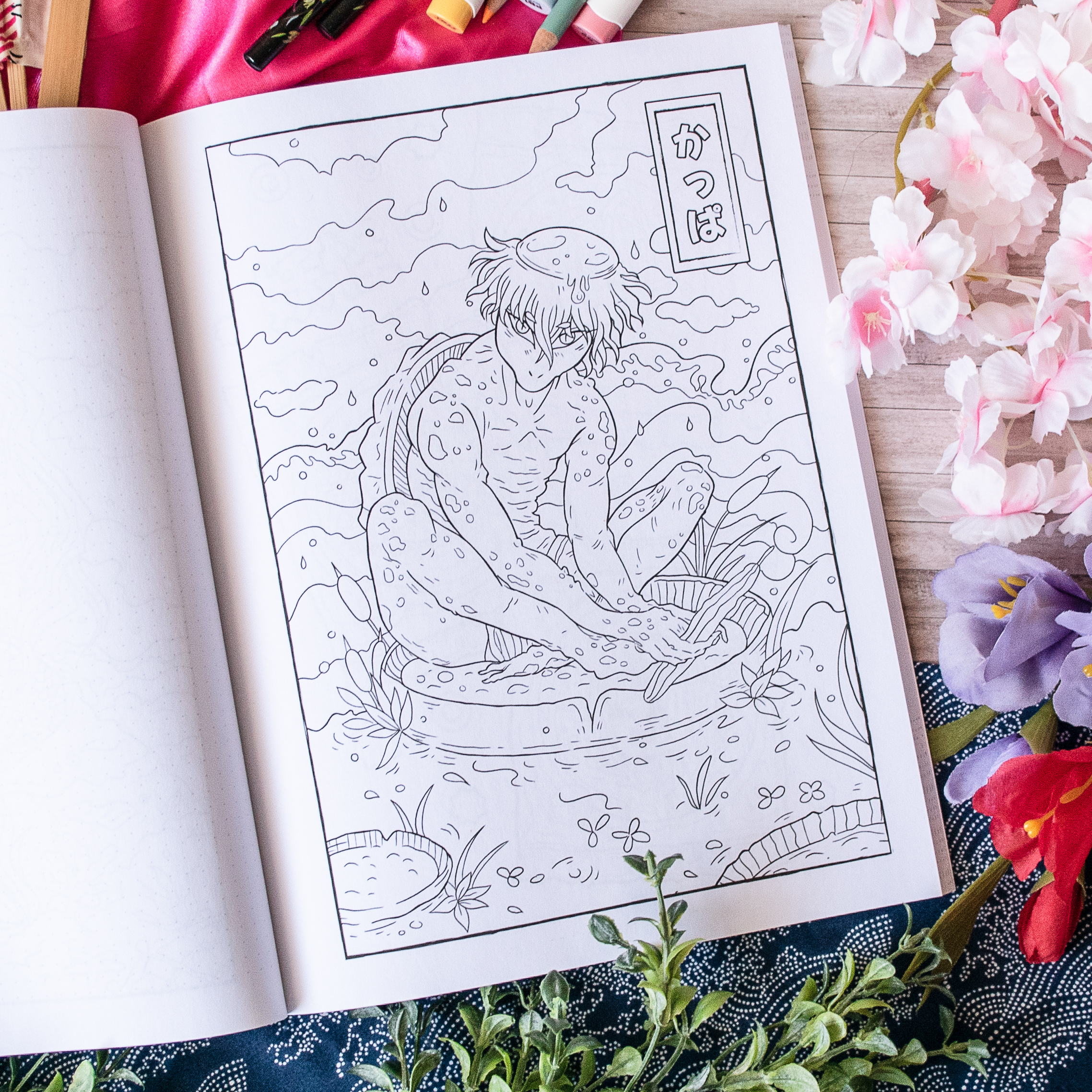 Une page tirée de « Coloriages Folklore Japonais » représente un kappa assis sur un rocher au bord d'un étang, entouré d'eau, de plantes et de nuages, avec un texte en japonais dans un encadré en haut à droite. À proximité se trouvent du matériel de dessin et des fleurs.