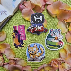Quatre autocollants d'animaux colorés sur un fond vert parsemé de pétales de rose roses : un chat en train de lire, un hamster dans une tasse à thé, un furet détective, ainsi qu'un crâne avec un os et un panda roux endormi.