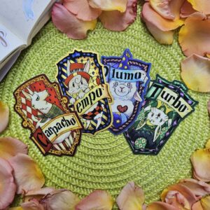 Quatre autocollants colorés représentant des animaux, intitulés « Carpacho », « Tempo », « Lumo » et « Turbo », sont disposés sur un tapis vert, entourés de pétales de roses roses et jaunes.