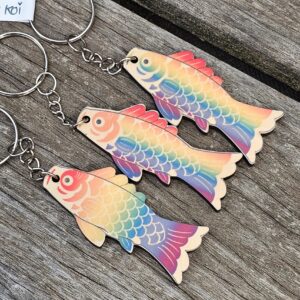 Trois porte-clés en bois aux couleurs de l'arc-en-ciel – « Queer Koi » – reposent sur une surface en bois patinée, disposés parallèlement les uns aux autres, avec des anneaux métalliques fixés à chaque poisson.