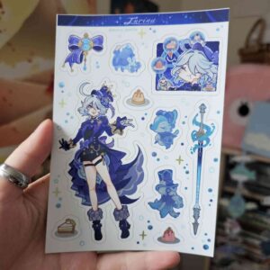 Une main tient l'autocollant « Kinich - Planche » au format A6, qui représente un personnage d'anime aux cheveux bleus vêtu d'une tenue élégante, entouré d'accessoires thématiques et d'adorables créatures bleues. L'arrière-plan est flou.