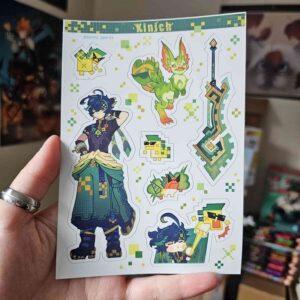 Une main tient la feuille d’autocollants « Kinich - Planche Stickers format A6 », sur laquelle figurent un personnage en pixel art, un dragon vert, une épée et plusieurs petits animaux. La feuille est bordée d’une frise pixelisée vert-jaune et porte l’inscription « Kinich » en haut.