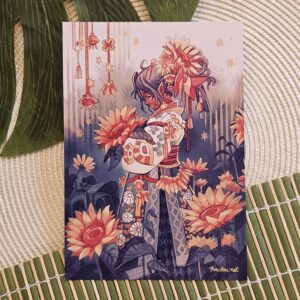 Une carte illustrée représente un personnage vêtu d'un habit richement orné de motifs, entouré de grands tournesols et d'ornements suspendus, dans une ambiance onirique rehaussée de touches étoilées. La carte repose sur une surface tissée, avec des feuilles vertes à proximité.