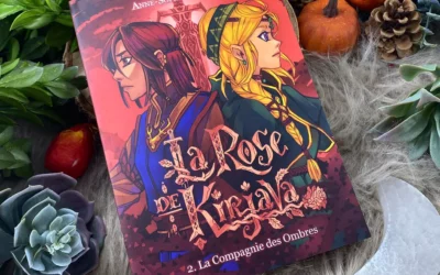 Un roman graphique intitulé *La Rose de Kirjava • Tome 2* repose sur de la fausse fourrure, entouré de plantes grasses, de pives, de citrouilles et de feuilles vertes. La couverture représente deux personnages illustrés dos à dos.