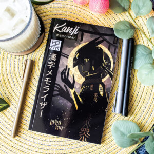 Un livre pour mémoriser les kanji, dont la couverture noire et dorée représente une silhouette stylisée allongée sur une natte tissée, entourée de stylos, d'une boisson et de feuilles vertes.