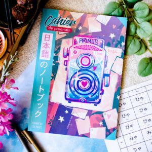 Un cahier de japonais au format A4 aux couleurs vives, intitulé « Cahier de japonais : Je révise avec Sophie 先生 Sensei (120 pages) », dont la couverture est ornée d’une illustration représentant un appareil photo rétro entouré de plantes, d’un stylo et d’une fiche d’exercices sur les hiragana, avec des textes en japonais et en français.