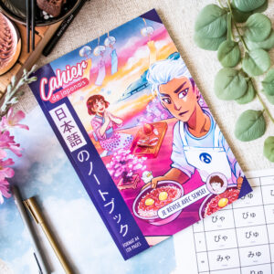 Cahier d'exercices de japonais aux couleurs vives, avec une couverture illustrée représentant deux personnages de style anime en train de manger. Ils sont entourés d'articles de papeterie, de plantes et d'une feuille d'exercices comportant des caractères hiragana écrits à la main. Le titre comporte la mention « Cahier de japonais » ainsi qu'un texte en japonais.