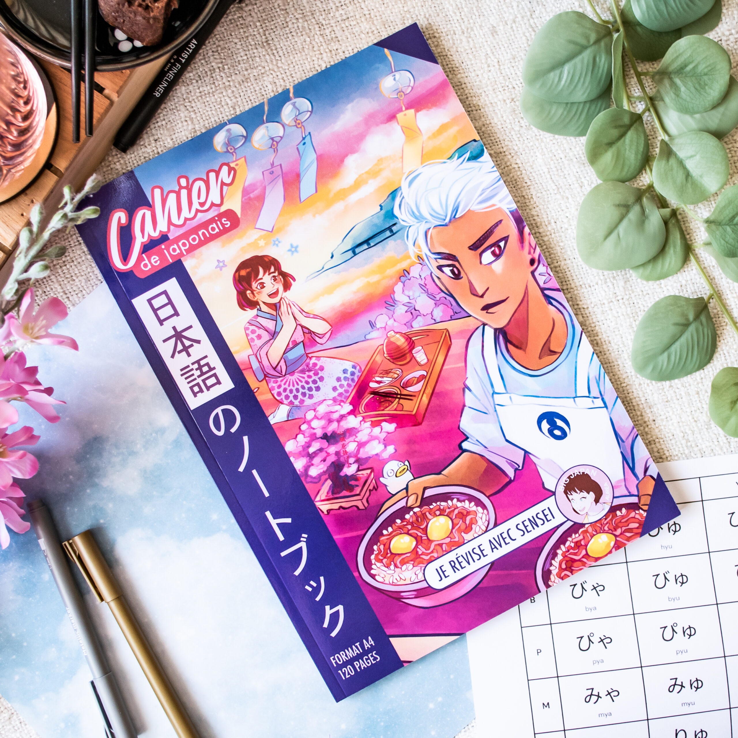 Cahier d'exercices de japonais aux couleurs vives, avec une couverture illustrée représentant deux personnages de style anime en train de manger. Ils sont entourés d'articles de papeterie, de plantes et d'une feuille d'exercices comportant des caractères hiragana écrits à la main. Le titre comporte la mention « Cahier de japonais » ainsi qu'un texte en japonais.