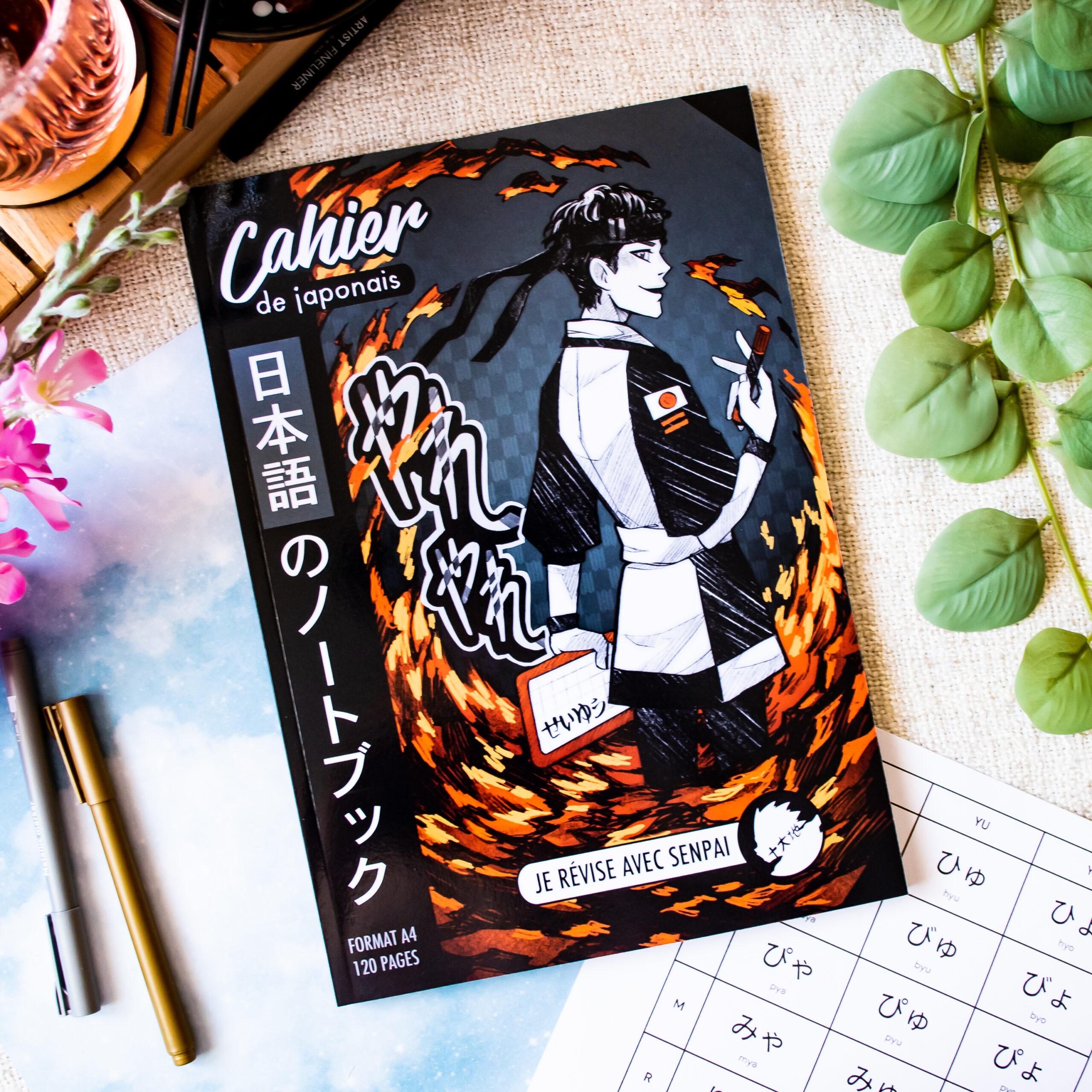 Voici la phrase révisée en fonction des informations fournies sur votre produit : Le « Cahier de japonais : Je révise avec Sophie 先生 Sensei » (format A4, 120 pages) présente une couverture de style anime représentant un personnage en noir et blanc entouré de flammes orange, posé sur une table aux côtés de stylos, d'une feuille d'exercices et de fleurs.
