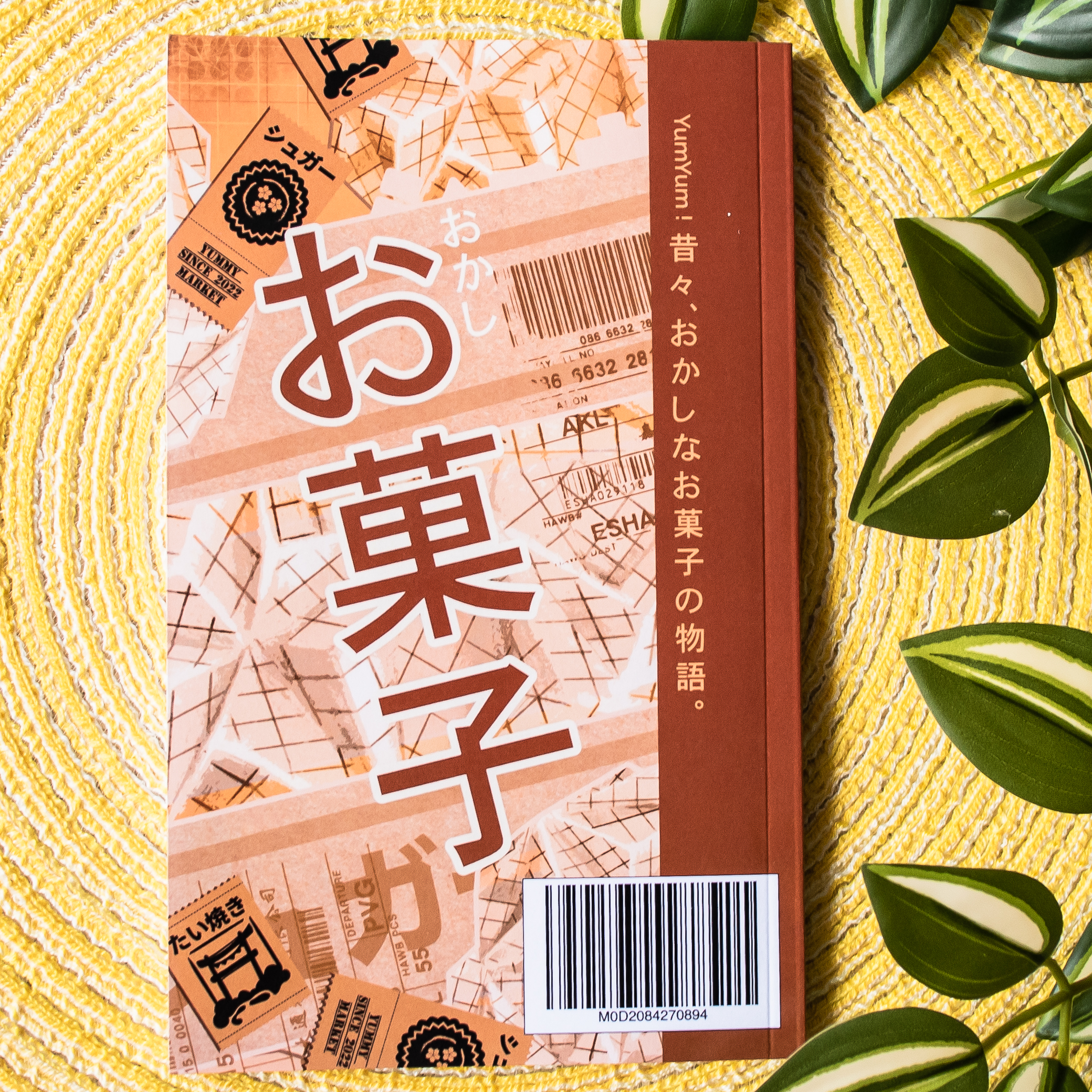 Un livre consacré aux emballages de snacks japonais repose sur une natte tissée jaune, avec des feuilles vertes à proximité. La couverture arbore des caractères japonais en gras, des codes-barres et des motifs décoratifs dans des tons de marron et de pêche.