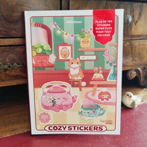 Un livre d'autocollants coloré intitulé « Cozy Stickers » présente sur sa couverture d'adorables animaux de dessin animé, des théières et des plantes, le tout dans une pièce chaleureuse. Une étiquette rouge indique : « Plus de 700 autocollants super mignons pour tout décorer... ».