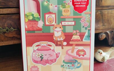 Un livre d'autocollants coloré intitulé « Cozy Stickers » présente sur sa couverture d'adorables animaux de dessin animé, des théières et des plantes, le tout dans une pièce chaleureuse. Une étiquette rouge indique : « Plus de 700 autocollants super mignons pour tout décorer... ».
