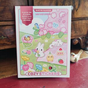 Un livre d'autocollants intitulé « Cozy Stickers », dont la couverture est ornée d'adorables illustrations représentant un lapin, un poussin, un gâteau, une fraise et des fleurs. Le livre est posé sur une surface en bois, à côté d'une petite figurine d'oiseau.